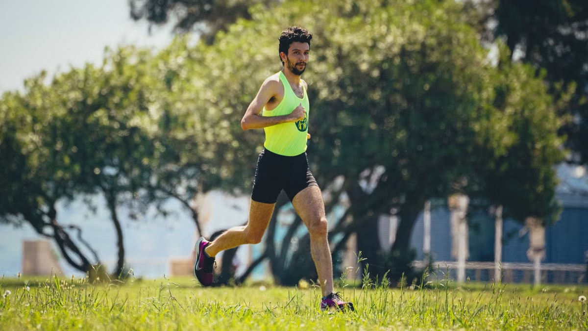 Full length of young bearded sportsman in activewear running on grassy lawn in city park near green trees during outdoor workout
